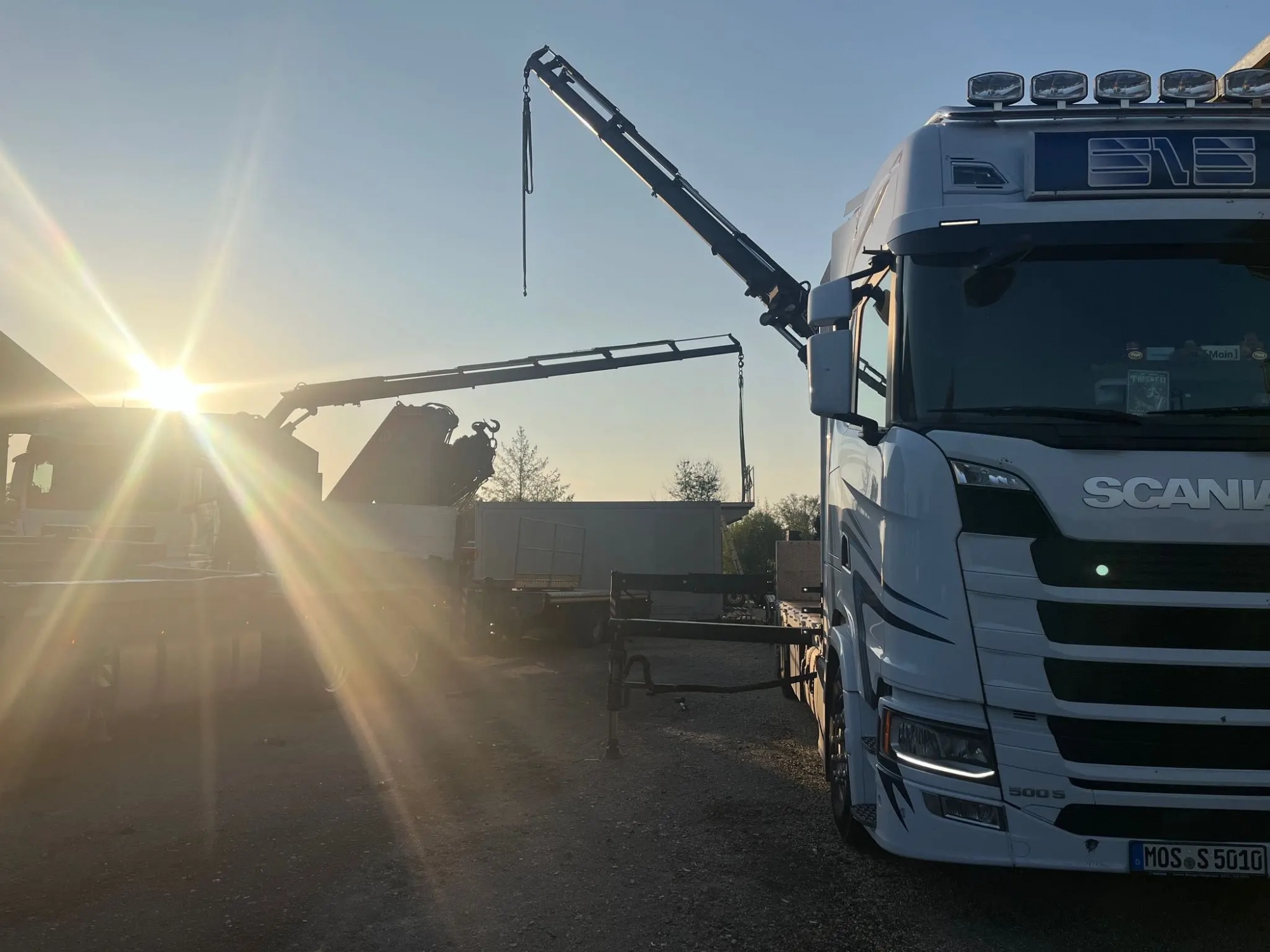 551b52ba-e86d-4fec-a765-32a9a8fb5099 Kran-LKW von SLS hebt einen Container bei Sonnenuntergang auf eine Baustelle